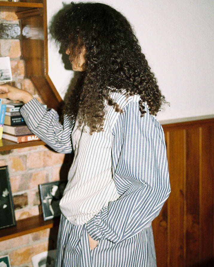 A person with long, curly dark hair wearing a blue and white vertically striped button-up shirt and matching striped pants, standing in a room with a wooden bookshelf and brick wall.