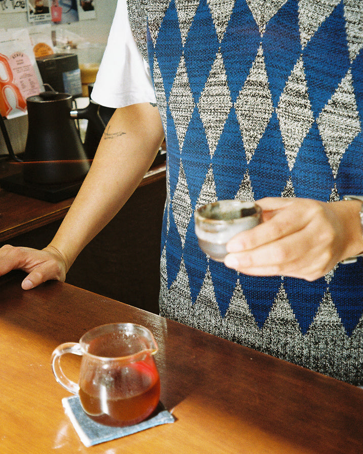 A person wearing a blue, gray, and white argyle sweater vest over a white t-shirt, holding a small glass cup on a wooden counter next to a glass pitcher with amber liquid.
