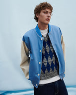 A man wears a blue varsity jacket over a diamond jacquard vest and dark blue jeans against a blue backdrop.