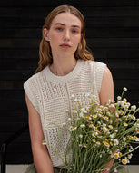 A women sits in front of a black weatherboard background wearing a cream knitted vest and green shorts holding a bunch of daisys.