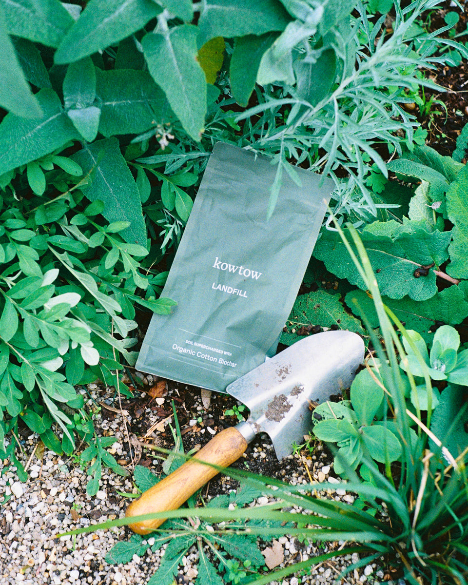 A matte dark green coffee bean bag labeled 'KOW KOW LANDSCAPE' lies on the ground among green plants, with a small hand trowel placed beside it.