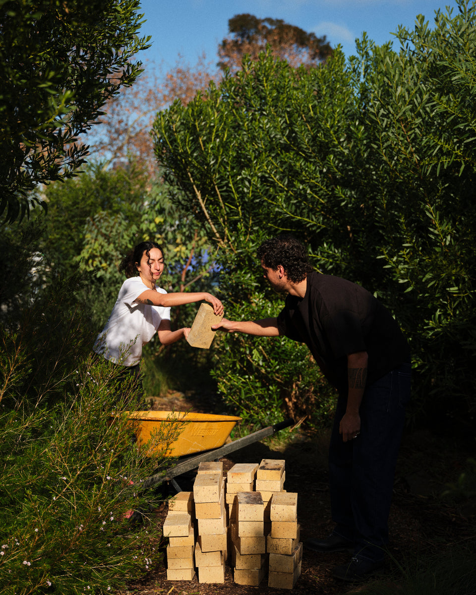 Two people outdoors in a garden or orchard, handing over rectangular clay or brick-like tiles. A yellow wheelbarrow sits behind them, and a stack of similar tiles is piled in the foreground among dense green plants under bright sunlight.