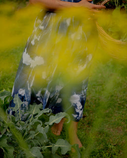 A person standing barefoot in a grassy outdoor garden, wearing a flowing blue and white tie-dye dress and holding a woven basket, with bright yellow flowers in the foreground.