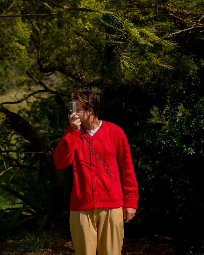 A person standing outdoors in a wooded area, wearing a bright red button up cardigan and beige pants, holding a camera to their face.