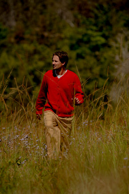 A man wearing a red knit cardigan and tan pants walking through a grassy outdoor field with trees in the background.