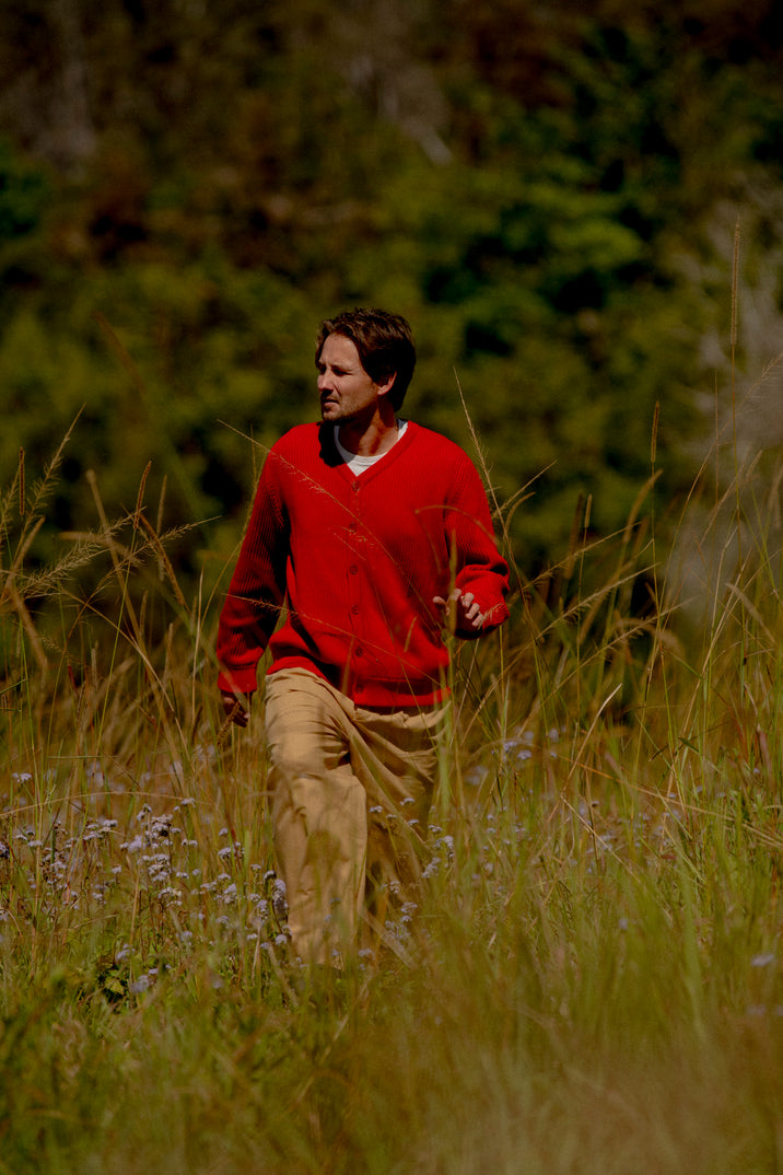 A man wearing a red knit cardigan and tan pants walking through a grassy outdoor field with trees in the background.
