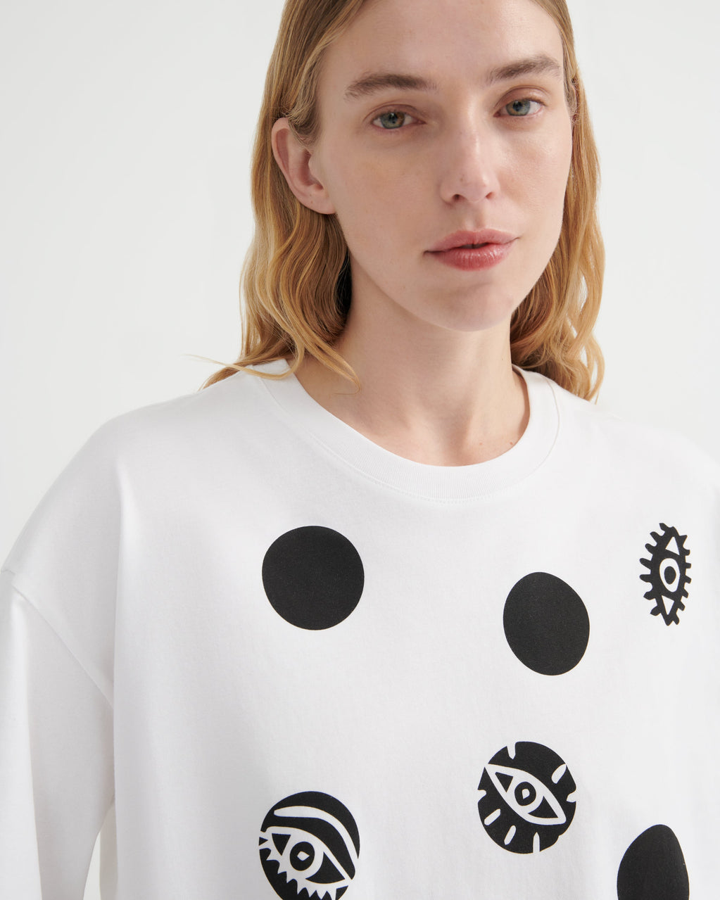 A close-up of a model wearing a white crewneck sweatshirt with black graphic prints, including solid circles and stylized eyes, against a plain light background.