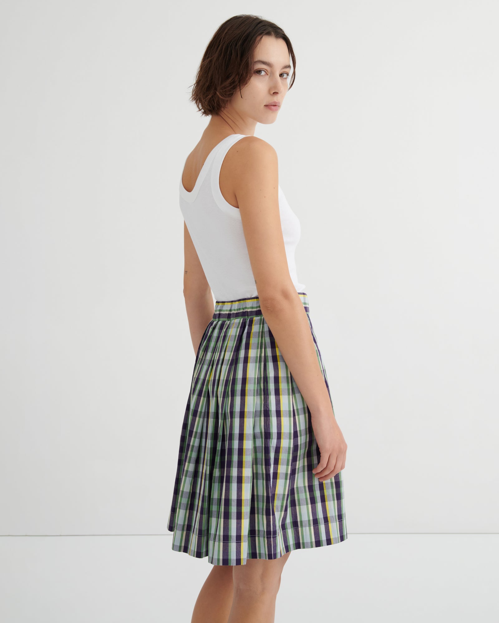 A woman wearing a white ribbed tank top and a green, navy, and yellow plaid pleated skirt, photographed from the back against a plain white studio background.