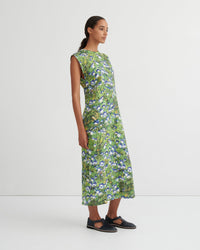 A woman modeling a sleeveless, knee-length green dress with a dense blue and white floral print, paired with black Mary Jane flats, against a plain white studio background.