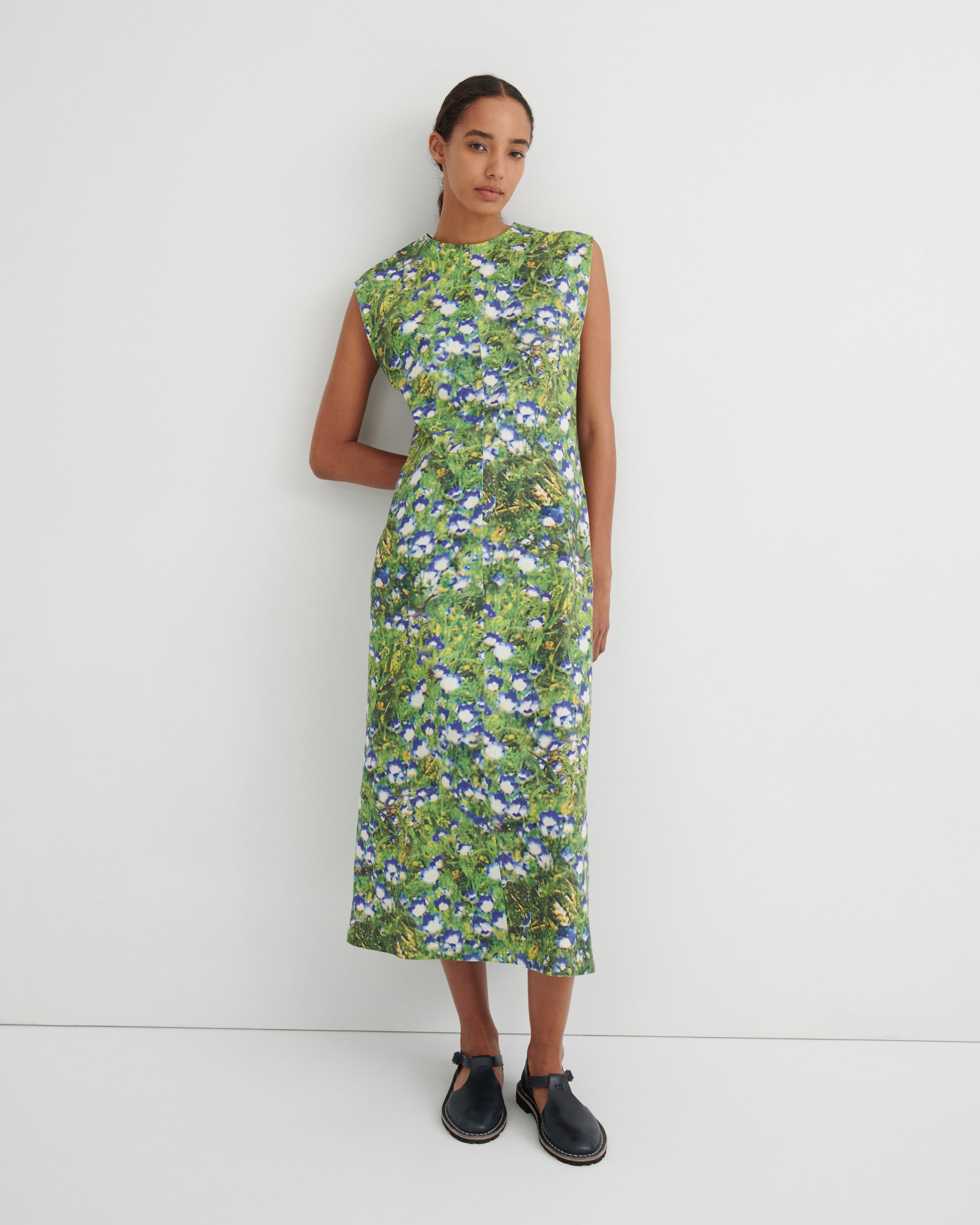 A woman modeling a sleeveless midi dress with a green and blue floral print, standing against a plain white studio background.