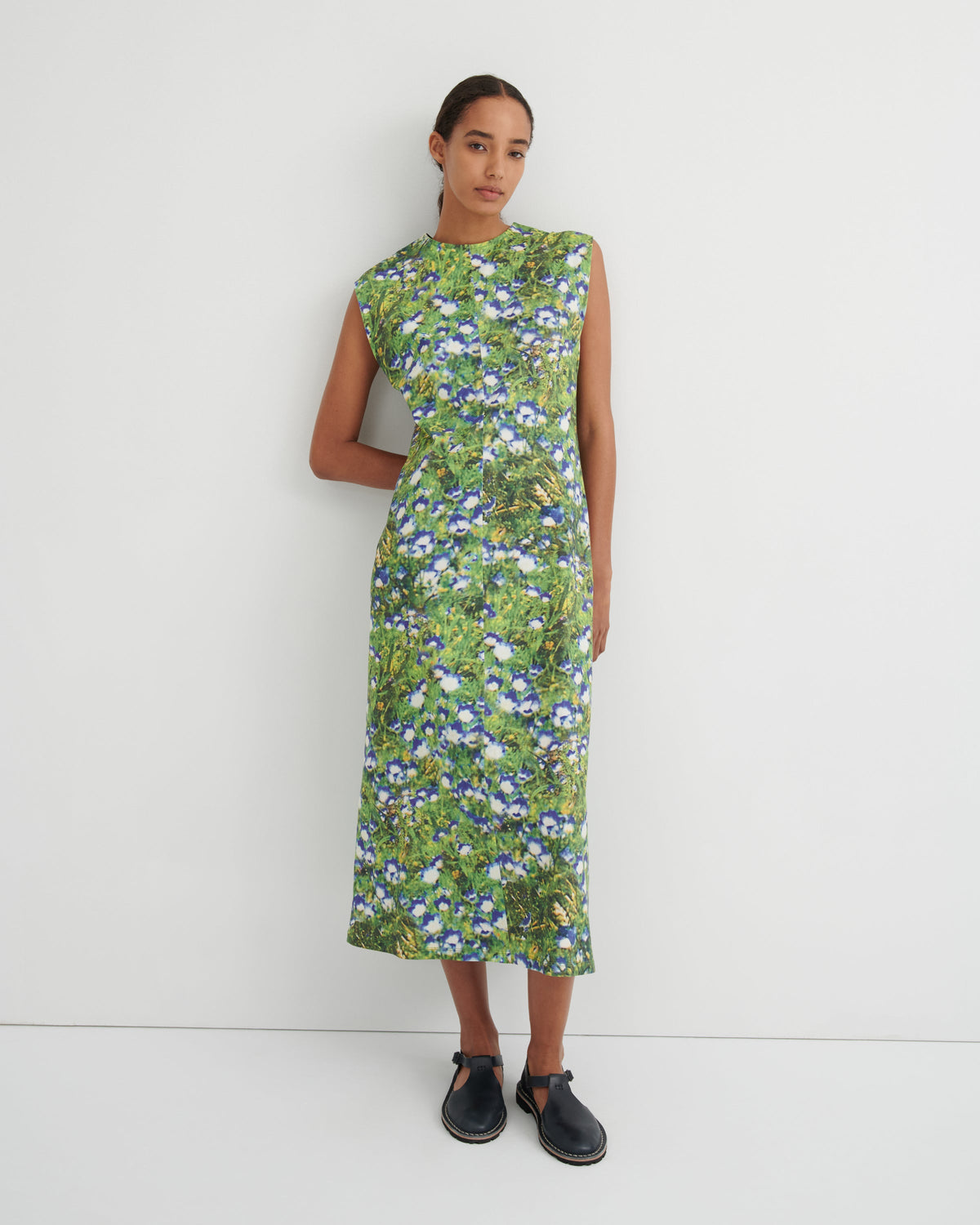 A woman modeling a sleeveless midi dress with a green and blue floral print, standing against a plain white studio background.
