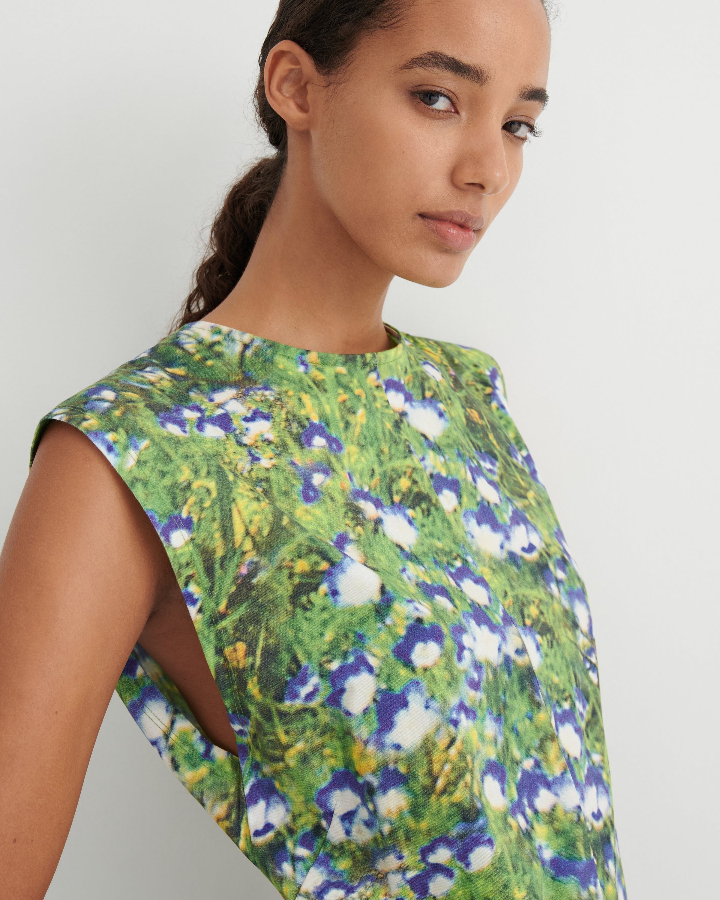 A close-up of a model wearing a sleeveless top with a vibrant green, blue, and white floral print, set against a plain light gray background.