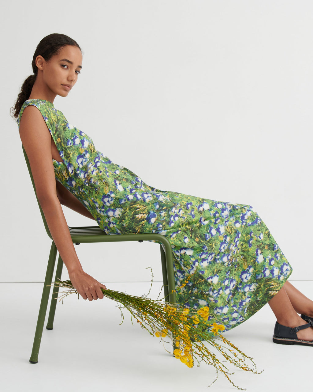A woman sits on a green metal chair wearing a sleeveless, knee-length green floral dress with blue and yellow flowers. She is holding a bouquet of yellow wildflowers against a plain white studio background.