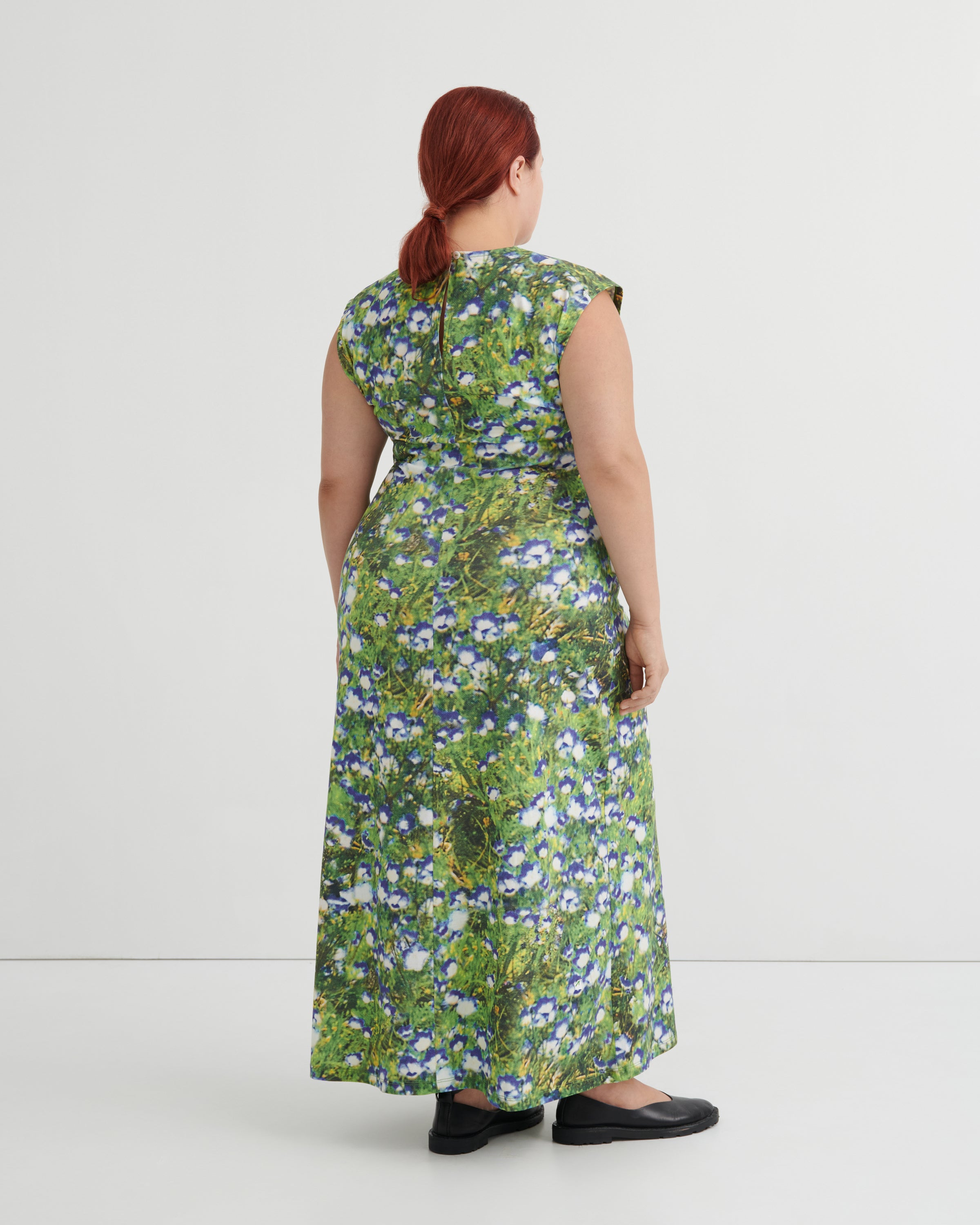 A woman with red hair tied back is shown from behind wearing a sleeveless, floor-length green dress with a dense blue and white floral print, paired with black flat shoes, against a plain light gray studio background.