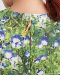 Close-up back view of a person wearing a floral print top with green foliage, purple flowers, and white blossoms, featuring a central seam and a small white button at the neckline.