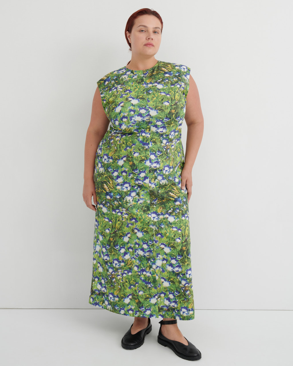A woman modeling a sleeveless, knee-length maxi dress with a green and blue floral print, paired with black flat shoes, against a plain light gray studio background.