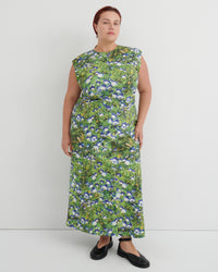 A woman modeling a sleeveless, knee-length maxi dress with a green and blue floral print, paired with black flat shoes, against a plain light gray studio background.