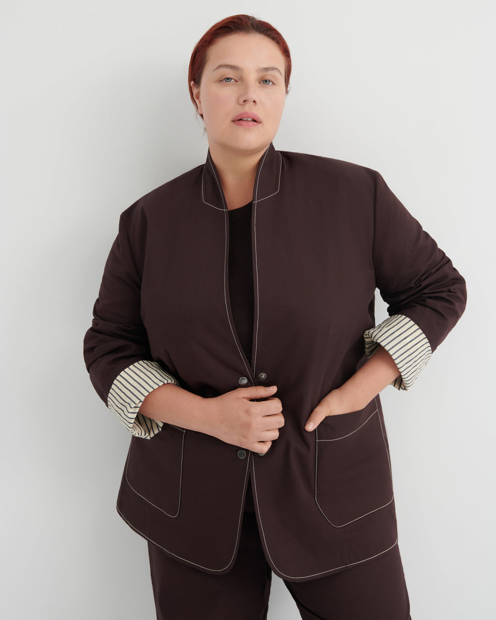 A woman modeling a dark brown jacket with contrasting white topstitching and striped button-up cuffs, paired with matching dark brown trousers against a light gray studio background.