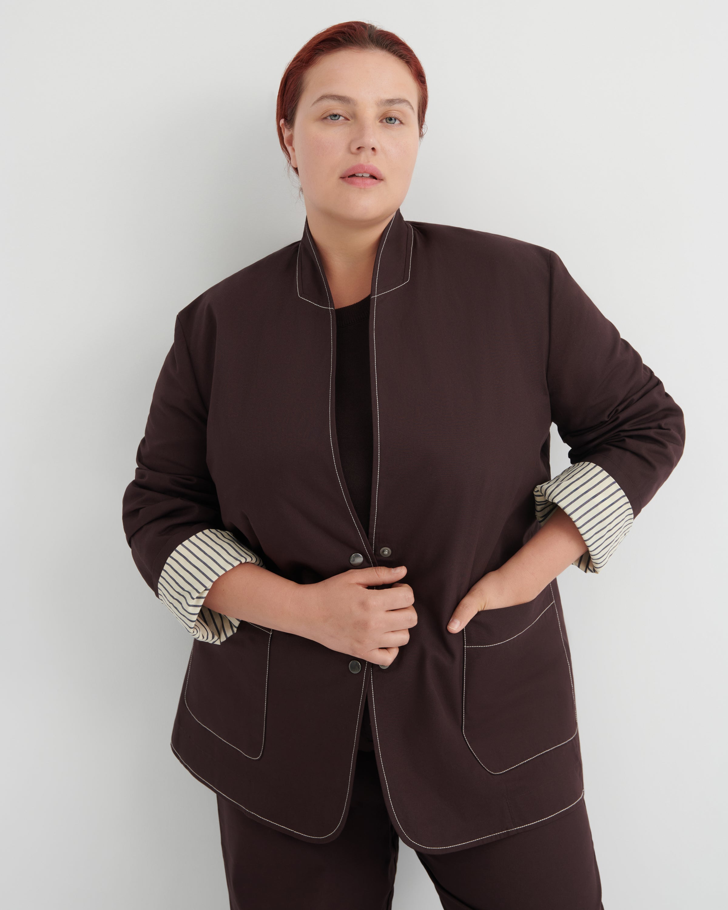 A woman modeling a dark brown jacket with contrasting white topstitching and striped button-up cuffs, paired with matching dark brown trousers against a light gray studio background.