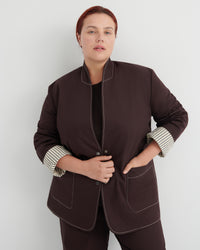 A woman modeling a dark brown jacket with contrasting white topstitching and striped button-up cuffs, paired with matching dark brown trousers against a light gray studio background.