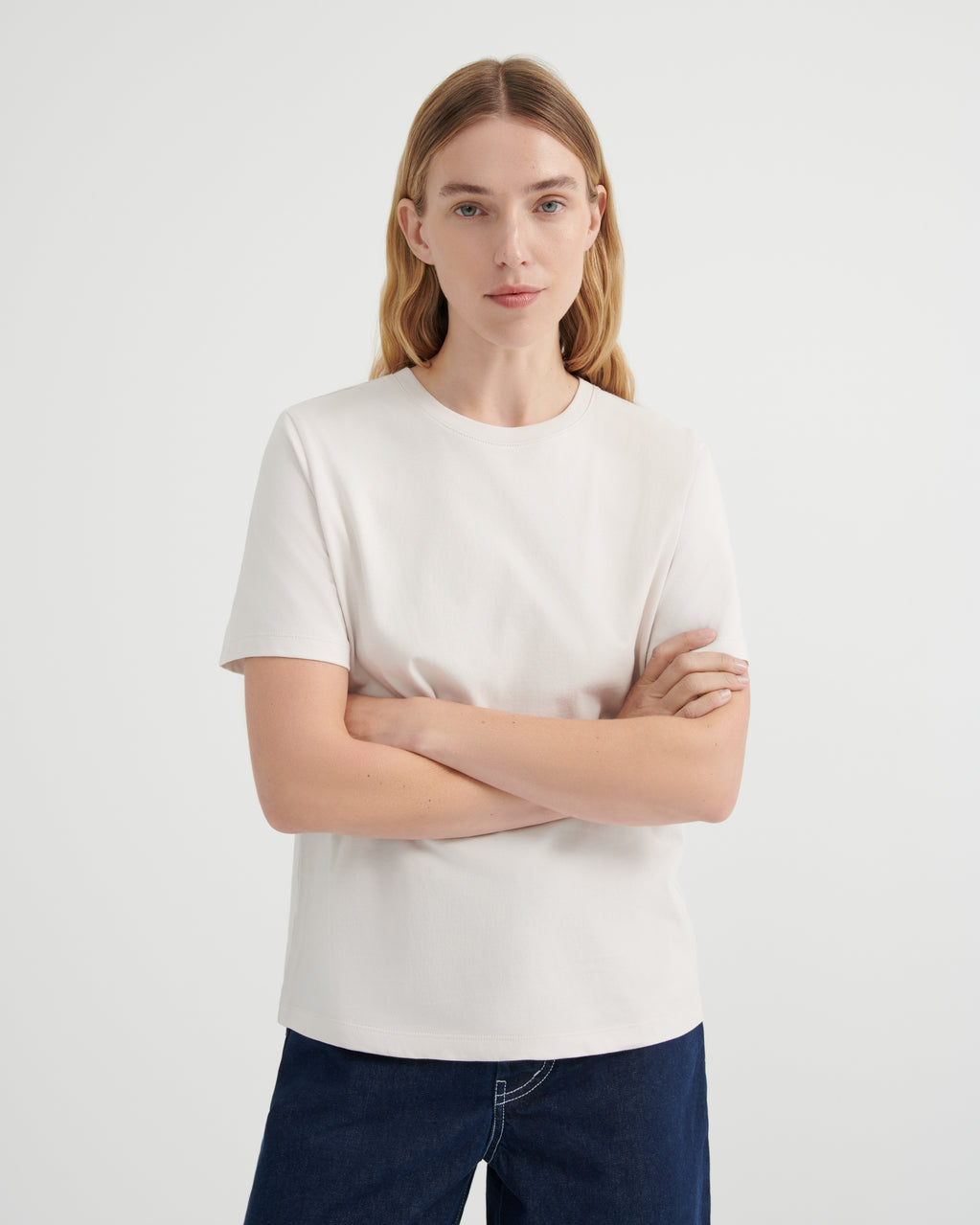 A woman with blonde hair wearing a plain off-white short-sleeve crewneck t-shirt and dark blue jeans, standing against a light gray studio background with her arms crossed.