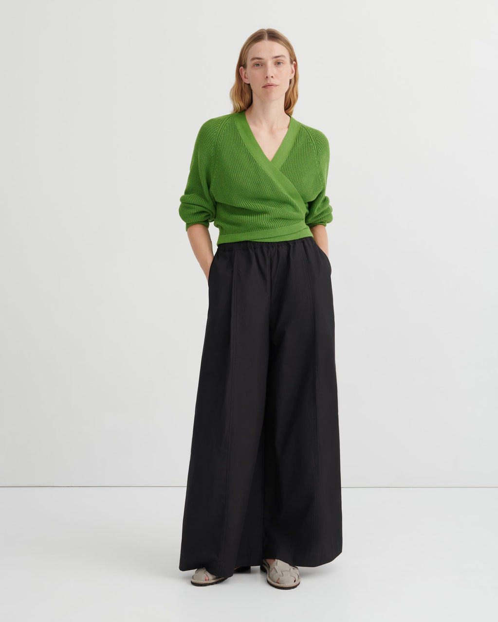 A woman stands against a plain light gray studio background wearing a bright green wrap-style V-neck sweater and wide-leg black trousers, with gray slip-on shoes.