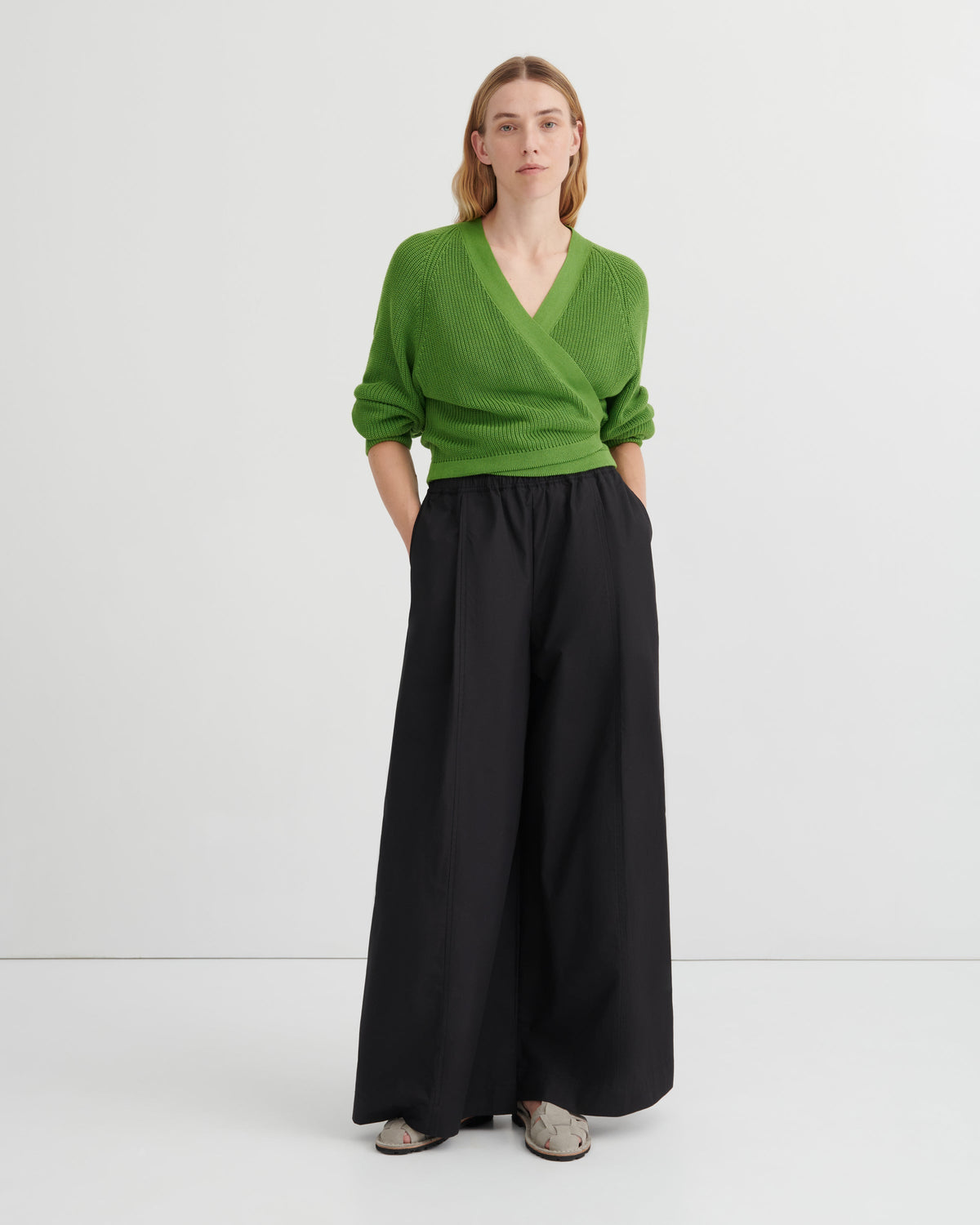 A woman stands against a plain light gray studio background wearing a bright green wrap-style V-neck sweater and wide-leg black trousers, with gray slip-on shoes.