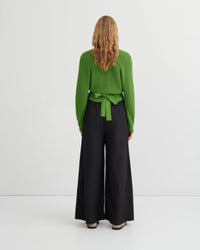 A woman viewed from the back wearing a bright green long-sleeve knit top with a tied waist and black wide-leg trousers, standing against a plain light gray studio background.