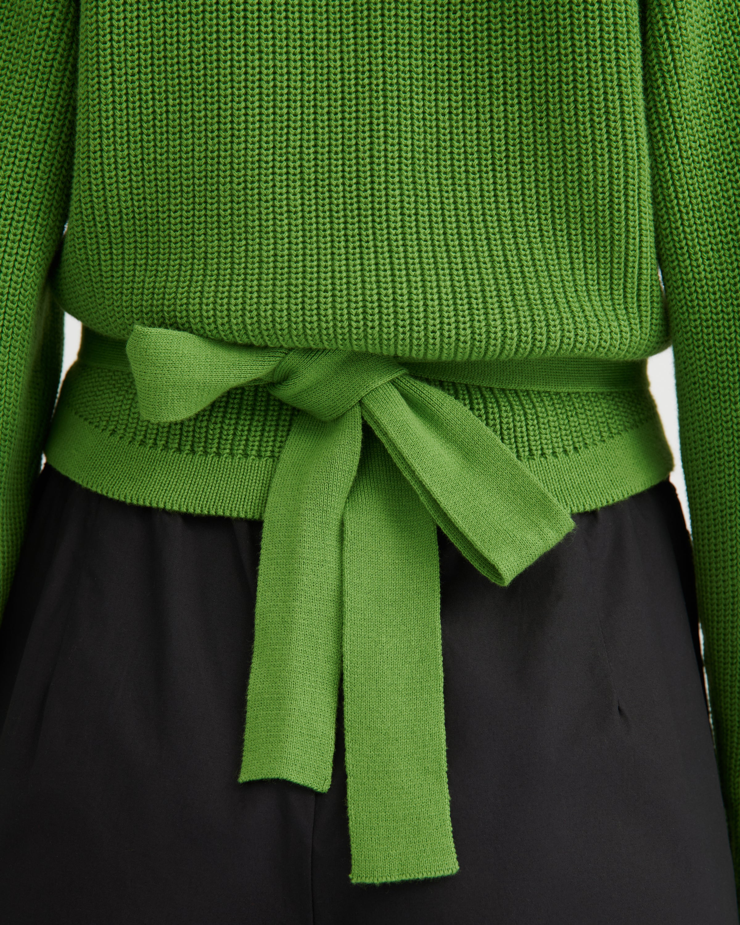 Close-up of a bright green ribbed knit sweater tied at the waist with a matching fabric belt, paired with black bottoms.