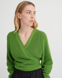 A woman wearing a bright green ribbed knit wrap sweater with a V-neck and long sleeves, paired with a black skirt, photographed against a light gray studio background.