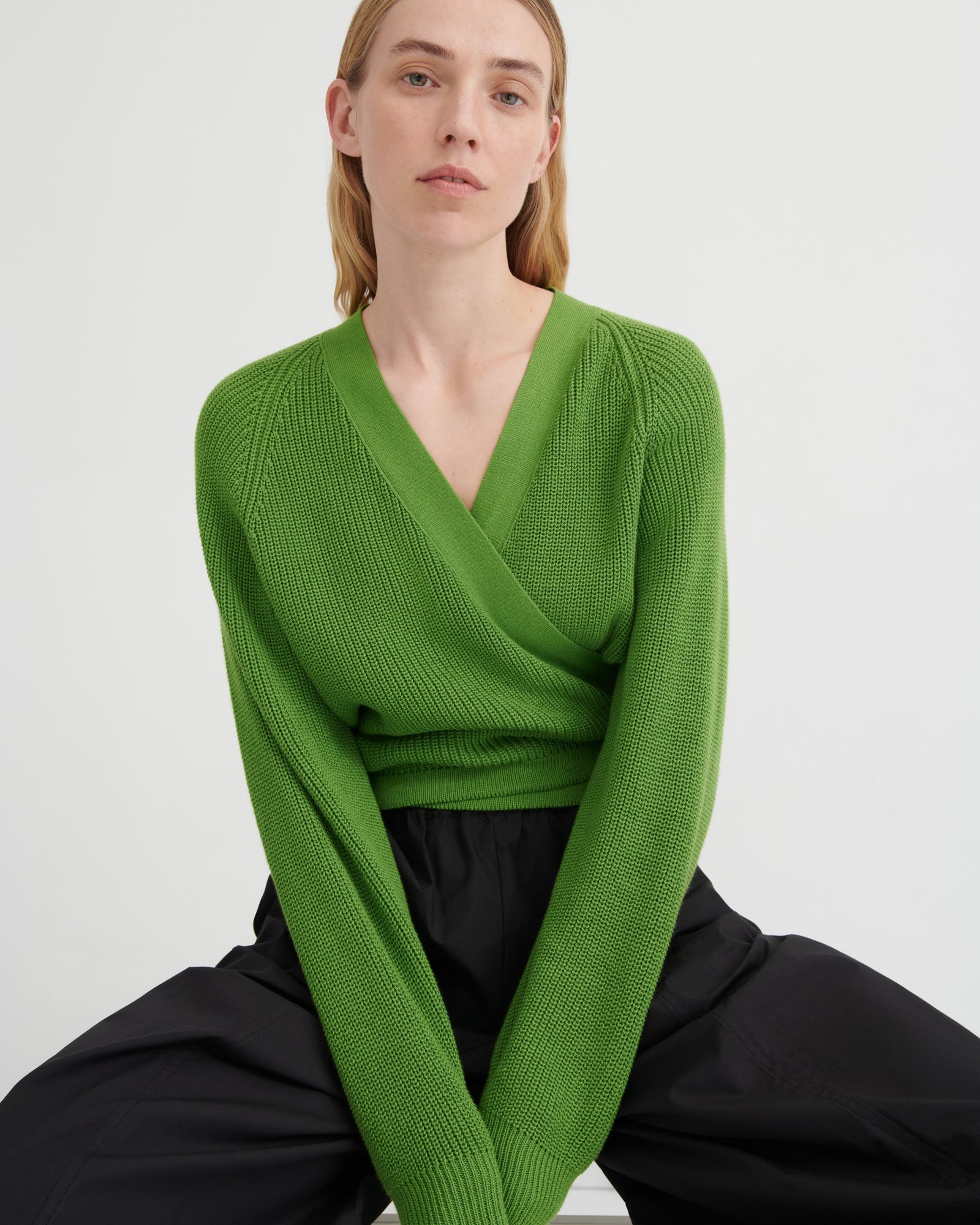 A woman wearing a bright green ribbed knit wrap sweater with long sleeves and a deep V-neck, paired with black pants, seated against a plain light gray background.