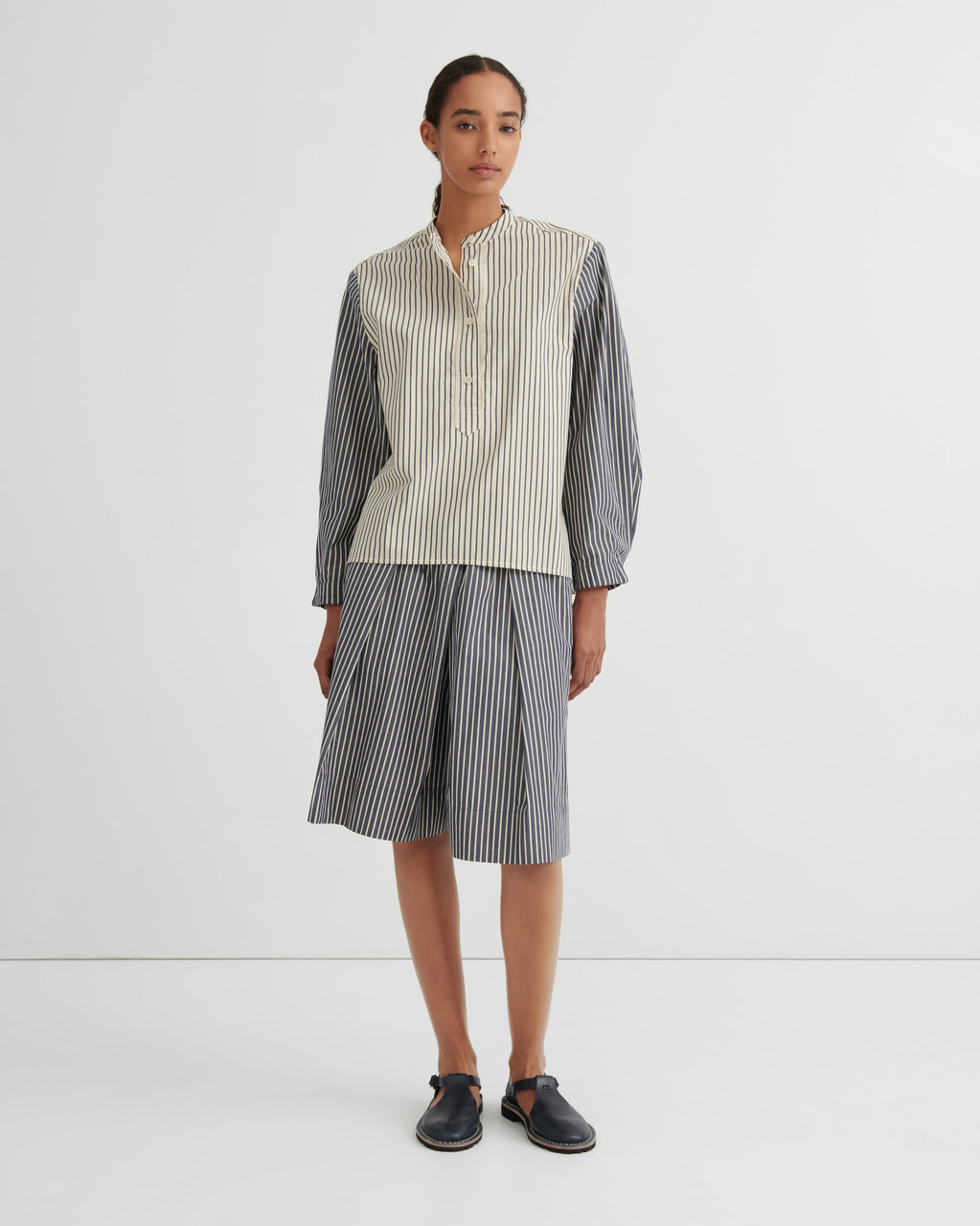 A model stands against a plain white studio background wearing a matching striped two-piece set consisting of a button up shirt and pleated shorts. The shirt has a cream and white vertical stripe pattern on the front and solid dark gray sleeves, while the shorts are entirely dark gray with thin white vertical stripes.