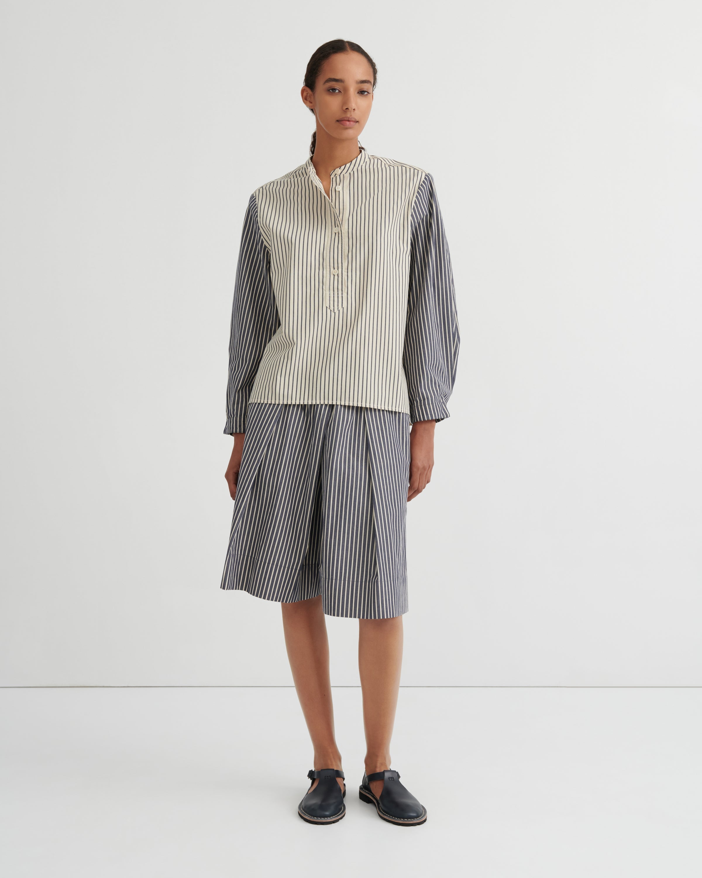 A model stands against a plain white studio background wearing a matching striped two-piece set consisting of a button up shirt and pleated shorts. The shirt has a cream and white vertical stripe pattern on the front and solid dark gray sleeves, while the shorts are entirely dark gray with thin white vertical stripes.