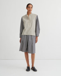 A model stands against a plain white studio background wearing a matching striped two-piece set consisting of a button up shirt and pleated shorts. The shirt has a cream and white vertical stripe pattern on the front and solid dark gray sleeves, while the shorts are entirely dark gray with thin white vertical stripes.