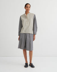 A model stands against a plain white studio background wearing a matching striped two-piece set consisting of a button up shirt and pleated shorts. The shirt has a cream and white vertical stripe pattern on the front and solid dark gray sleeves, while the shorts are entirely dark gray with thin white vertical stripes.