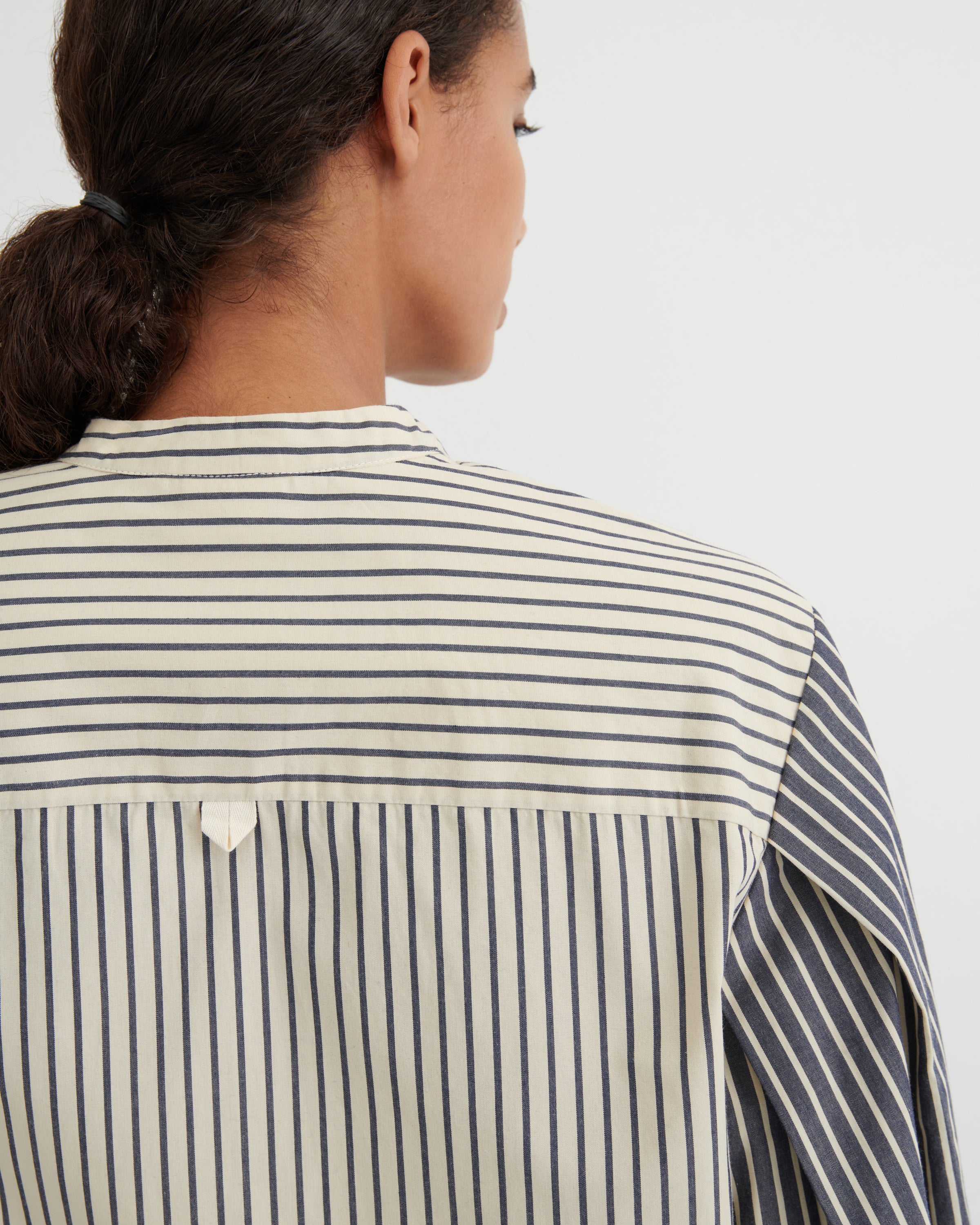 Close-up back view of a person wearing a striped shirt with horizontal and vertical stripe patterns in cream and dark navy, against a plain light gray background.
