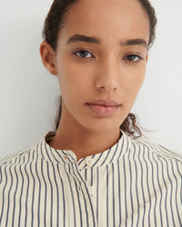 Close-up of a model wearing a cream-colored button-down shirt with thin navy vertical stripes, set against a plain light gray background.