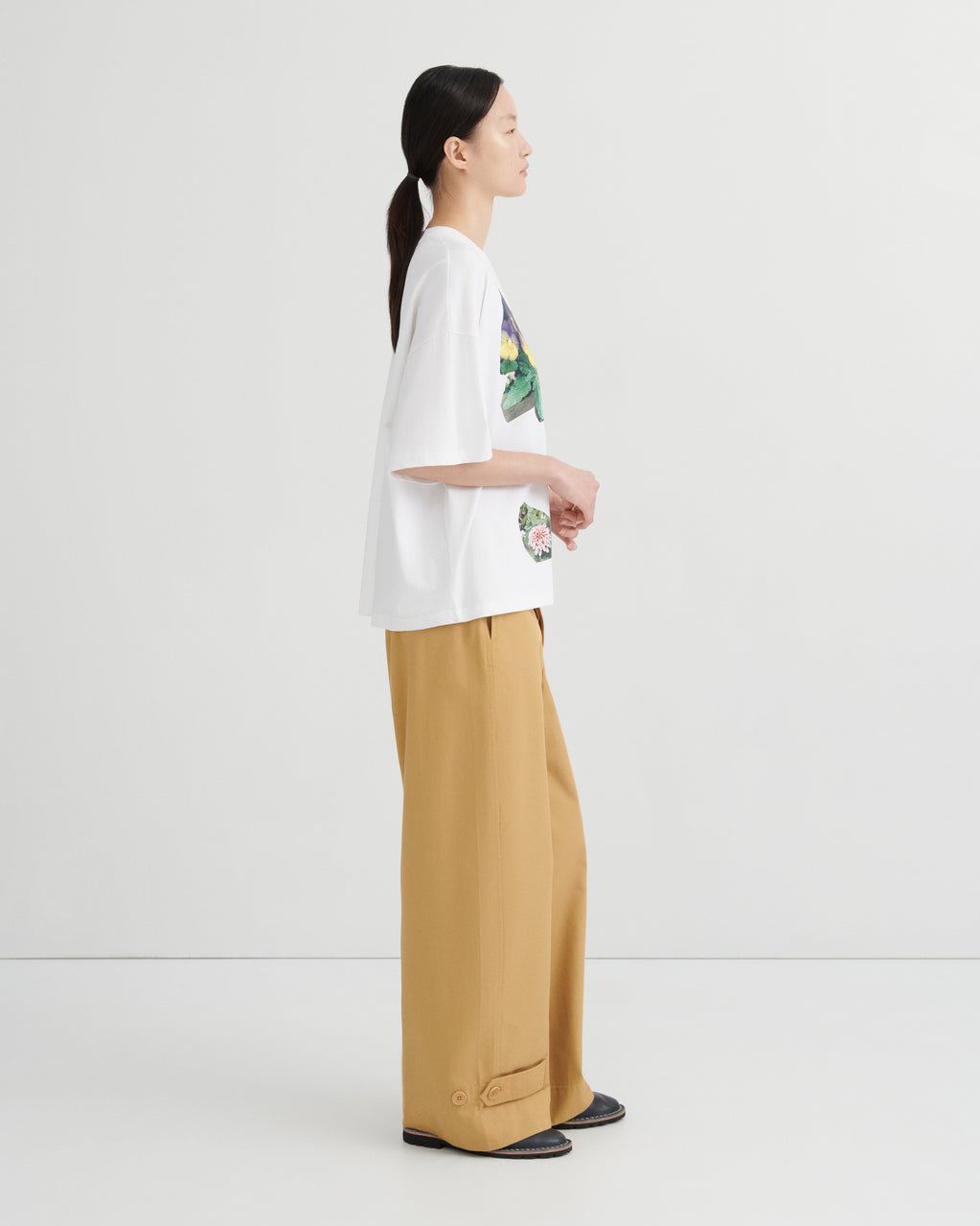 A woman shown in side profile wearing an oversized white graphic T-shirt and wide-leg tan pants against a clean light gray studio background.