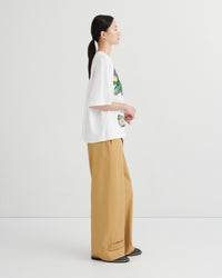 A woman shown in side profile wearing an oversized white graphic T-shirt and wide-leg tan pants against a clean light gray studio background.