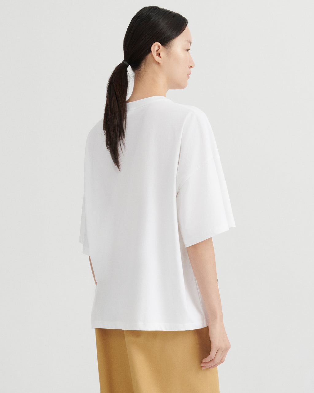 A woman shown from the back wearing an oversized white short-sleeve top and tan pants against a light gray studio background.