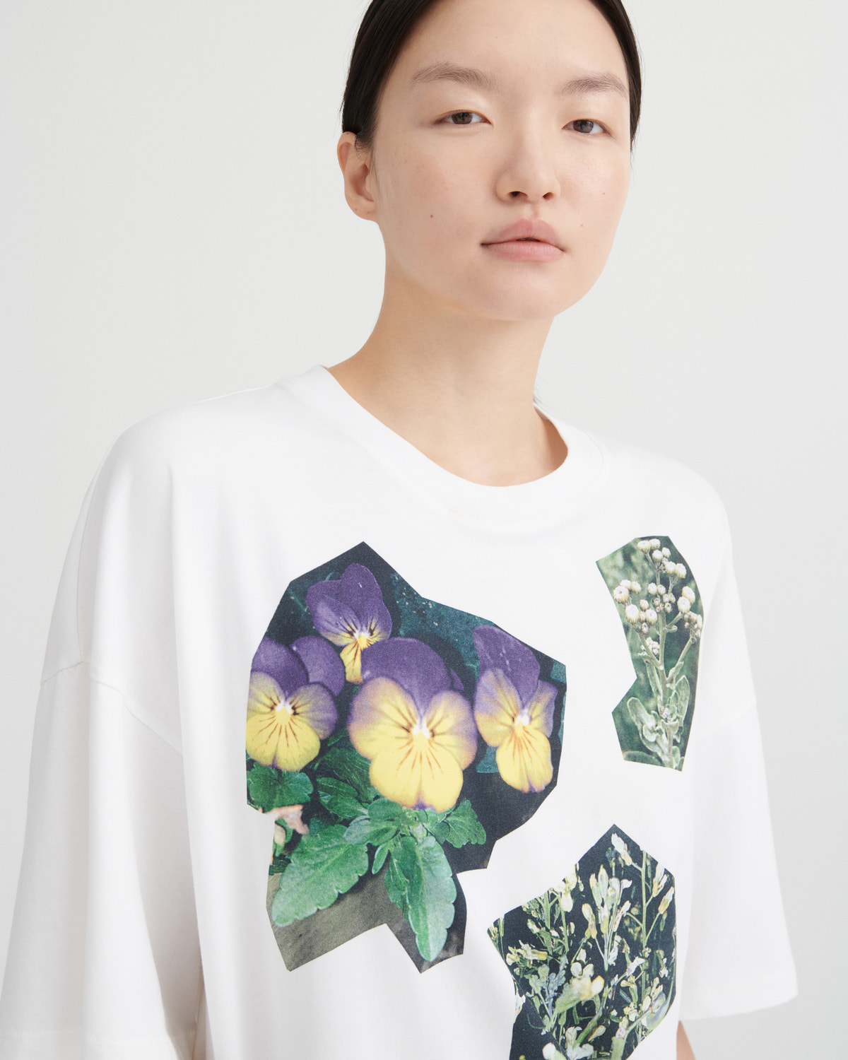 A model wearing a white oversized crewneck t-shirt featuring a graphic print of purple and yellow pansies and green botanical illustrations.