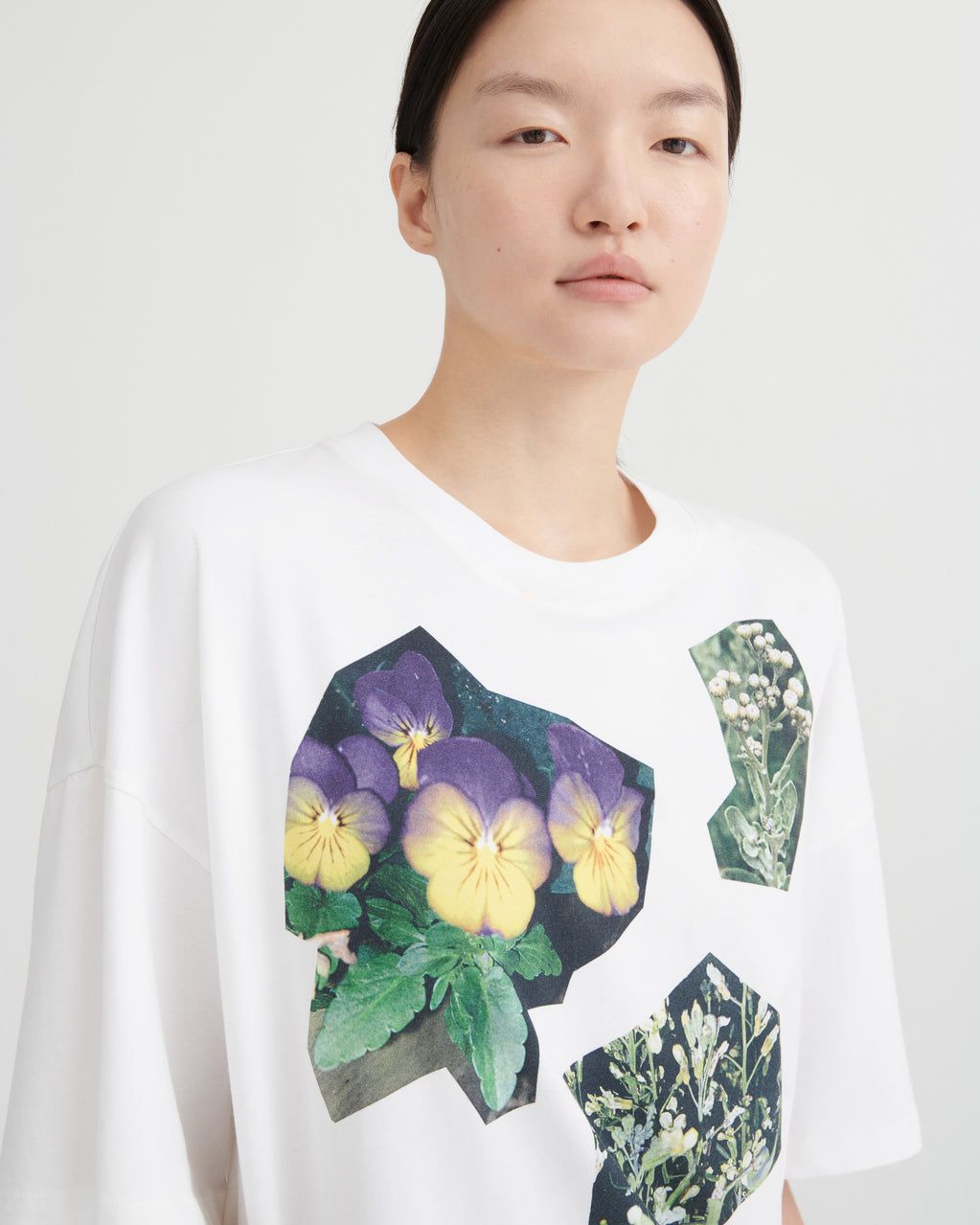 A model wearing a white oversized crewneck t-shirt featuring a graphic print of purple and yellow pansies and green botanical illustrations.