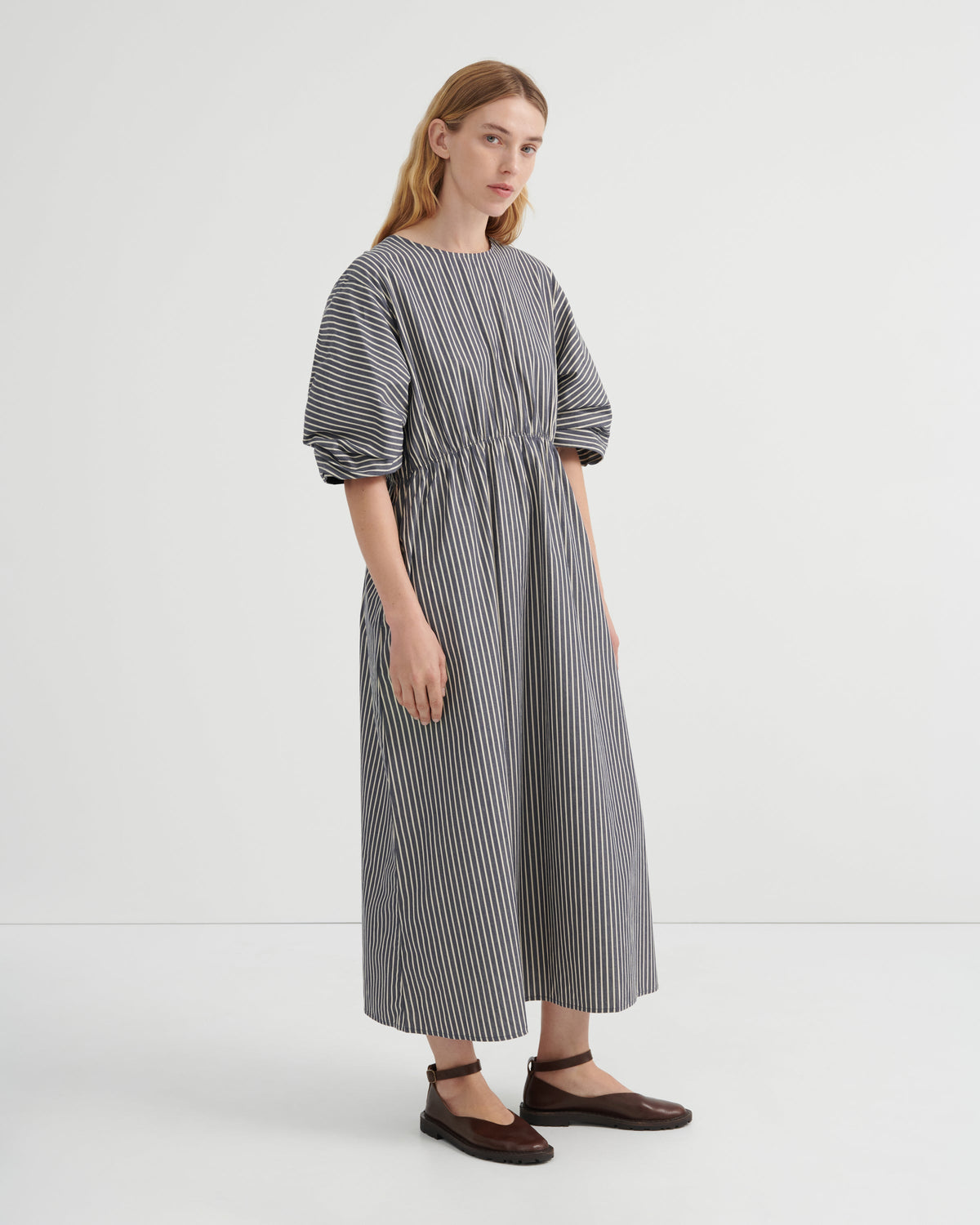 A woman modeling a loose-fitting midi dress with vertical black and white stripes, short puff sleeves, and a gathered waist, paired with dark brown flat shoes against a plain white studio background.