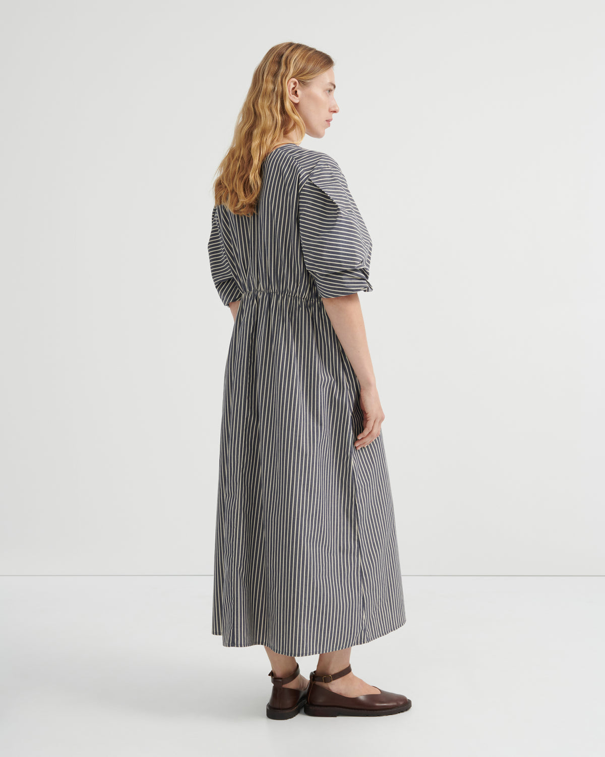 A woman modeling a long-sleeved midi dress with vertical black and white stripes, puff sleeves, and a gathered waist, shown from a side-back angle against a plain white studio background.