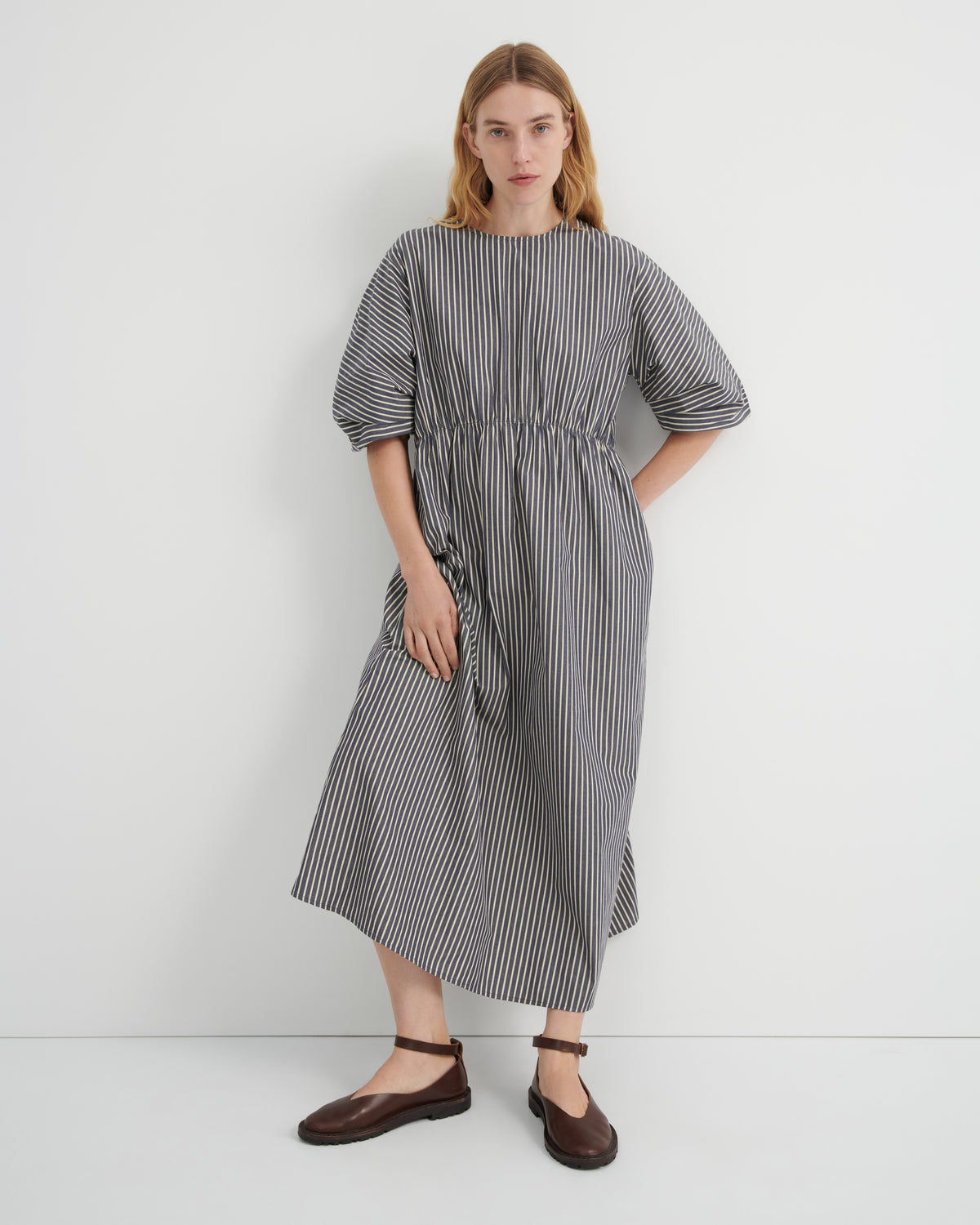 A woman modeling a loose-fitting midi dress with vertical black and white stripes, short puff sleeves, and a gathered waist, paired with dark brown flat shoes against a plain white studio background.