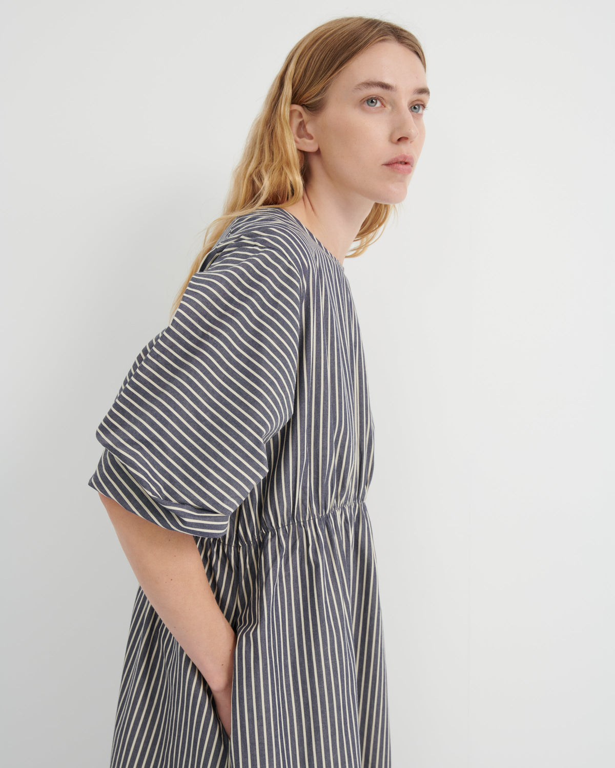 A woman wearing a loose-fitting, short-sleeved dress with vertical gray and white stripes, shown from a side profile against a plain light gray background.