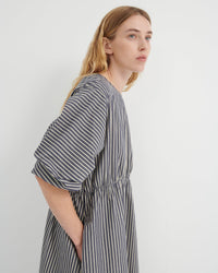 A woman wearing a loose-fitting, short-sleeved dress with vertical gray and white stripes, shown from a side profile against a plain light gray background.