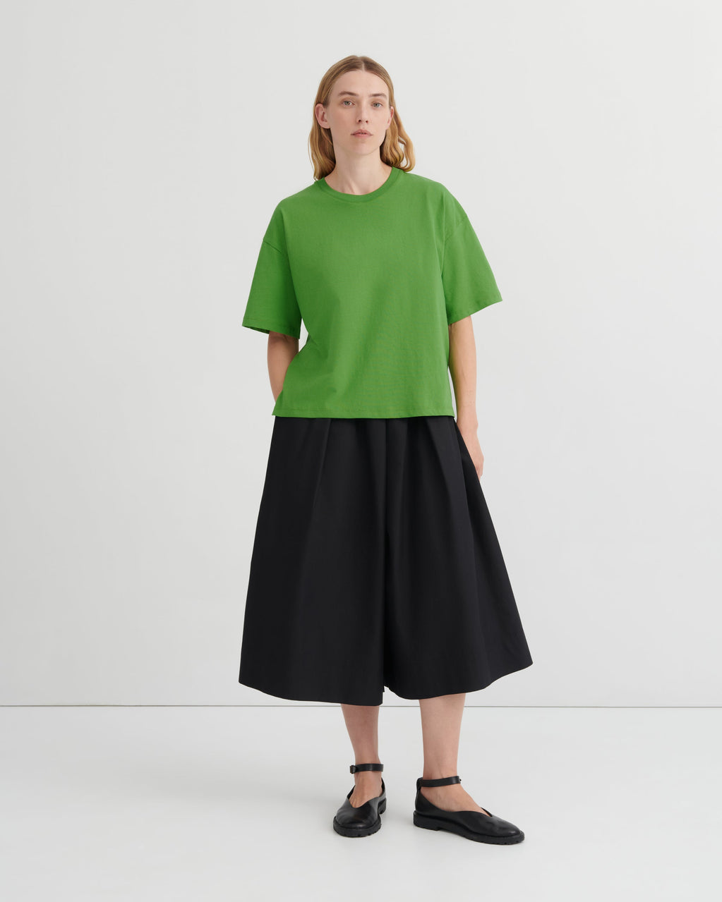 A model stands against a light gray studio background wearing a bright green short-sleeve crewneck top, a black pleated midi skirt, and black flat shoes.