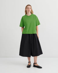 A model stands against a light gray studio background wearing a bright green short-sleeve crewneck top, a black pleated midi skirt, and black flat shoes.