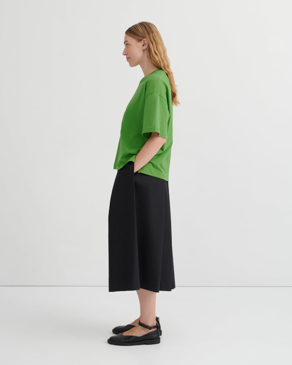 A woman stands in profile wearing an oversized bright green short-sleeve top, black wide-leg cropped pants, and black Mary Jane flats against a clean white studio background.